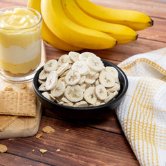 Dried banana chips in black bowl, fresh bananas, banana pudding, and graham crackers on wood table
