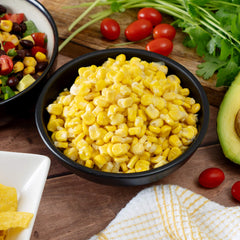 Bowl of sweet corn kernels on a wooden table with tomatoes, avocado, cilantro and salad