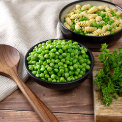 Fresh green peas in a bowl with pasta salad, parsley, and a wooden spoon on a table